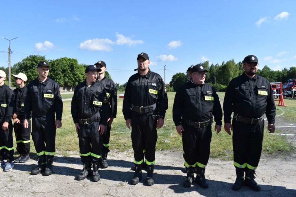 Na zewnątrz występuje grupa osób w mundurach stojących w szeregu na zewnątrz podczas promieni słonecznych dnia. Mają oni na sobie czarne mundury z neonowymi, żółtymi paskami przy sekcji części spodniej oraz czapki.