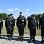 Na zewnątrz występuje grupa osób w mundurach stojących w szeregu na zewnątrz podczas promieni słonecznych dnia. Mają oni na sobie czarne mundury z neonowymi, żółtymi paskami przy sekcji części spodniej oraz czapki.