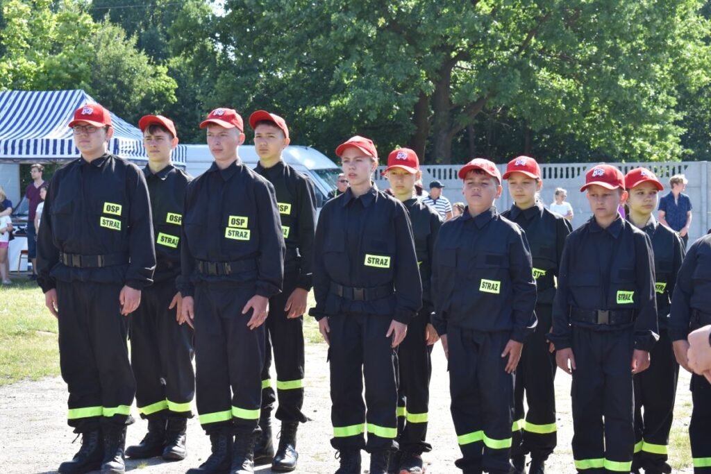 Na obrazku jest grupa umundurowanych osób ubranych w czarne stroje z odblaskowymi pasami i czerwonymi czapkami, stojącymi w szyku na świeżym powietrzu. W tle można dotrzeć na zewnątrz oraz namiot, co jest ważne, że scena odbywa się podczas różnych wydarzeń plenerowego.