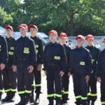 Na obrazku jest grupa umundurowanych osób ubranych w czarne stroje z odblaskowymi pasami i czerwonymi czapkami, stojącymi w szyku na świeżym powietrzu. W tle można dotrzeć na zewnątrz oraz namiot, co jest ważne, że scena odbywa się podczas różnych wydarzeń plenerowego.