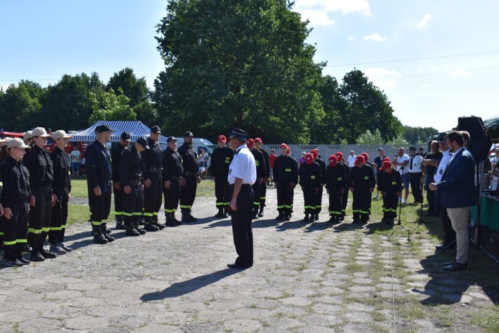 Na obrazku widnieje grupa umundurowanych osób, z których korzystają na głowach czerwonych czapki; wszystkie rozwiązania w szeregu na tle przyrody. W widocznych drzewach oraz ludzi ubranych w codziennych strojach.