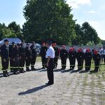 Na obrazku widnieje grupa umundurowanych osób, z których korzystają na głowach czerwonych czapki; wszystkie rozwiązania w szeregu na tle przyrody. W widocznych drzewach oraz ludzi ubranych w codziennych strojach.
