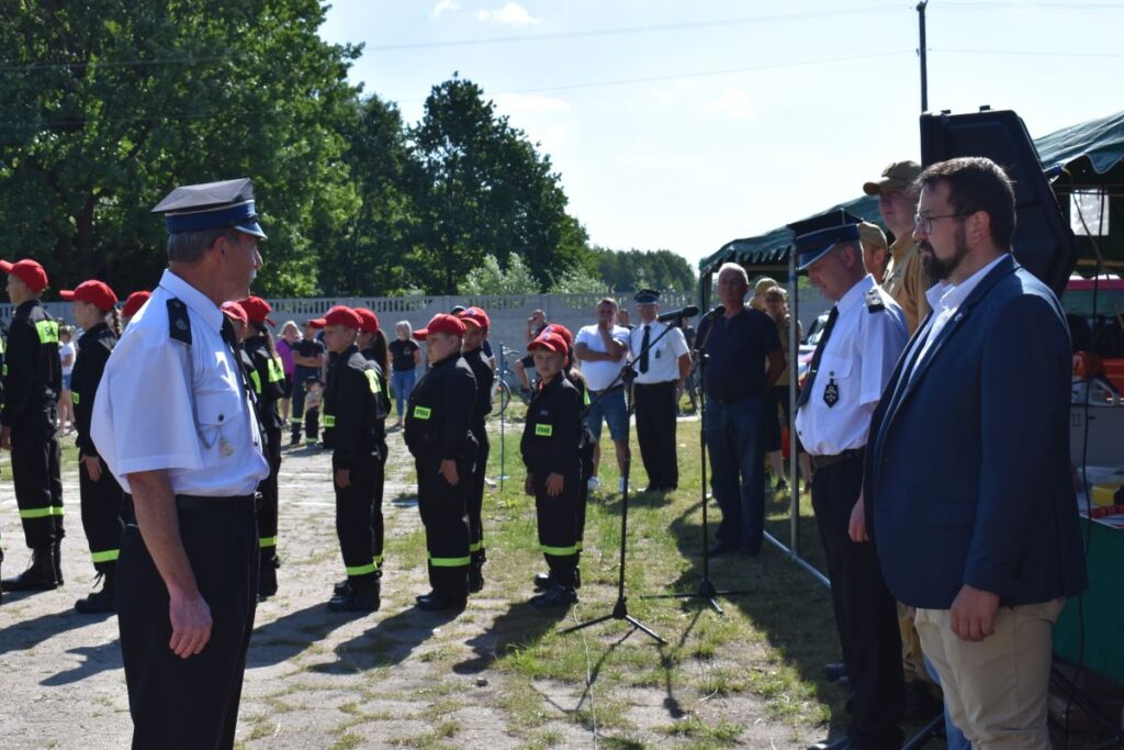 Na obrazku grupa umundurowanych osób stoi w formacji na świeżym powietrzu, zwrócona w stronę kilku oficjeli w odświętnych strojach. W tle można spędzić kilku obserwatorów oraz namiot, a wszystko na miejscu w słoneczny dzień.