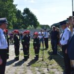 Na obrazku grupa umundurowanych osób stoi w formacji na świeżym powietrzu, zwrócona w stronę kilku oficjeli w odświętnych strojach. W tle można spędzić kilku obserwatorów oraz namiot, a wszystko na miejscu w słoneczny dzień.