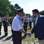Na obrazku osoby w koszuli mundurowej i czapce trzymanej przez inną osobę w ciemnym morzu podczas wydarzeń na świeżym powietrzu. W tle znajduje się osoba w mundurach i czerwonych czapkach ustalonych w szeregu.