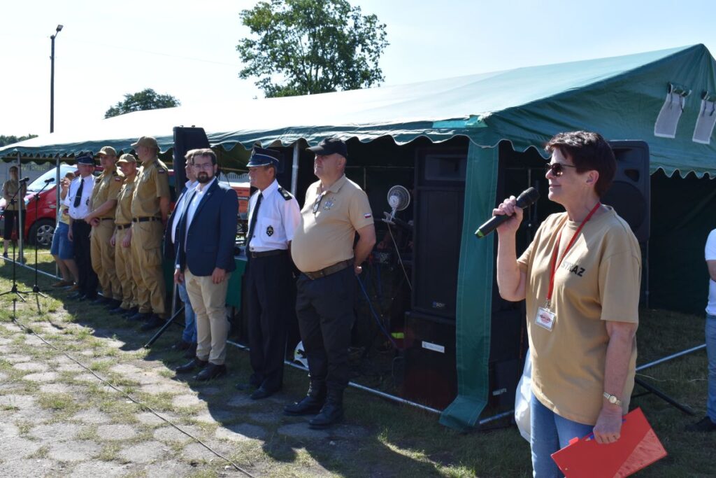 Na obrazku osoby z mikrofonem, która przemawia do grupy osób w mundurach, stojących przed zielonym namiotem podczas plenerowego wydarzeń. Tło umieszczone na zewnątrz, na zewnątrz, na świeżym powietrzu.