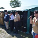Na obrazku osoby z mikrofonem, która przemawia do grupy osób w mundurach, stojących przed zielonym namiotem podczas plenerowego wydarzeń. Tło umieszczone na zewnątrz, na zewnątrz, na świeżym powietrzu.