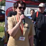 Na obrazie widać osobę w okularach graficznych i beżowej koszuli z napisem „STRAŻ”, która przemawia do mikrofonu podczas wydarzeń plenerowych. W tle znajdują się dwie inne osoby, z których jedna jest ubrana w czarnym mundurze i kapeluszu.