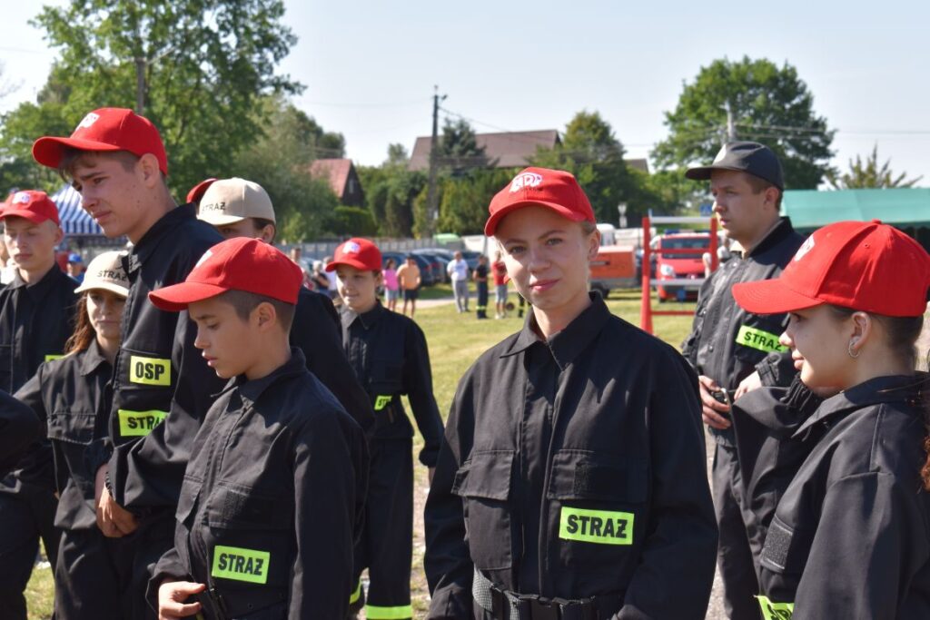 Zdjęcie przedstawia grupę ludzi w czarnych mundurach z naszymiywkami „STRAŻ” oraz czerwonemi czapkami, stojącymi na świeżym powietrzu w słoneczny dzień. W tle widoczne są różne drzewa i akcesoria.