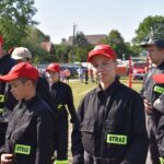 Zdjęcie przedstawia grupę ludzi w czarnych mundurach z naszymiywkami „STRAŻ” oraz czerwonemi czapkami, stojącymi na świeżym powietrzu w słoneczny dzień. W tle widoczne są różne drzewa i akcesoria.