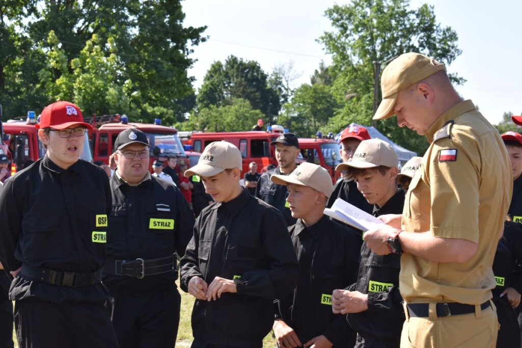 Na obrazku widnieje grupa młodych osób w czarnych mundurach i czapkach, słuchających mężczyzny w beżowym mundurze trzymającym notatnik. W tle kilka wozów strażackich emitowanych na słonecznym terenie.