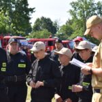 Na obrazku widnieje grupa młodych osób w czarnych mundurach i czapkach, słuchających mężczyzny w beżowym mundurze trzymającym notatnik. W tle kilka wozów strażackich emitowanych na słonecznym terenie.