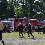 Na obrazku strażacy uczestniczyli w ćwiczeniach, symulując akcję niszczącą. W tle znajdują się kilka charakterystycznych wozów strażackich, a wszystko to, co jest przestrzegane przez zgromadzony tłum ludzi.
