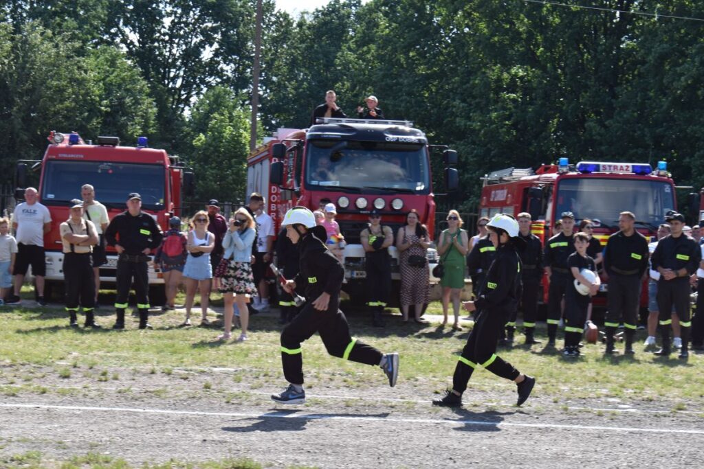 Trzech zawodników biegnie przez pole, na zasadzie spiesząc się do akcji ratunkowej. W tle widać wozy strażackie oraz tłum gapiów wykonawczych.