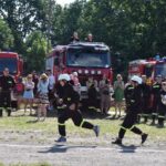 Trzech zawodników biegnie przez pole, na zasadzie spiesząc się do akcji ratunkowej. W tle widać wozy strażackie oraz tłum gapiów wykonawczych.