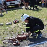 Na obrazku jednolitym w czarnym mundurze i białym hełmie zwijającym się na ziemi. W tle widoczny jest wóz strażacki oraz grupa występującychych osób.