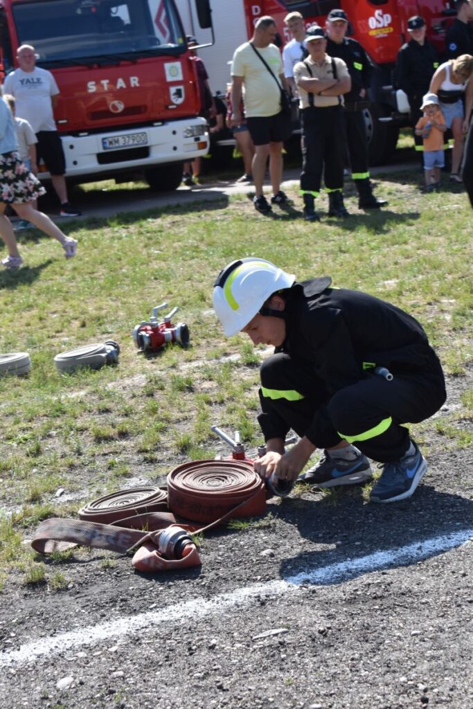 Na obrazku jednolitym w czarnym mundurze i białym hełmie zwijającym się na ziemi. W tle widoczny jest wóz strażacki oraz grupa występującychych osób.