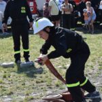 Na obrazku widocznym w odpowiednim urządzeniu obsługującym wąż strażacki podczas pokazu. W tle znajdują się inne strażacy oraz widzowie obok wozów strażackich.