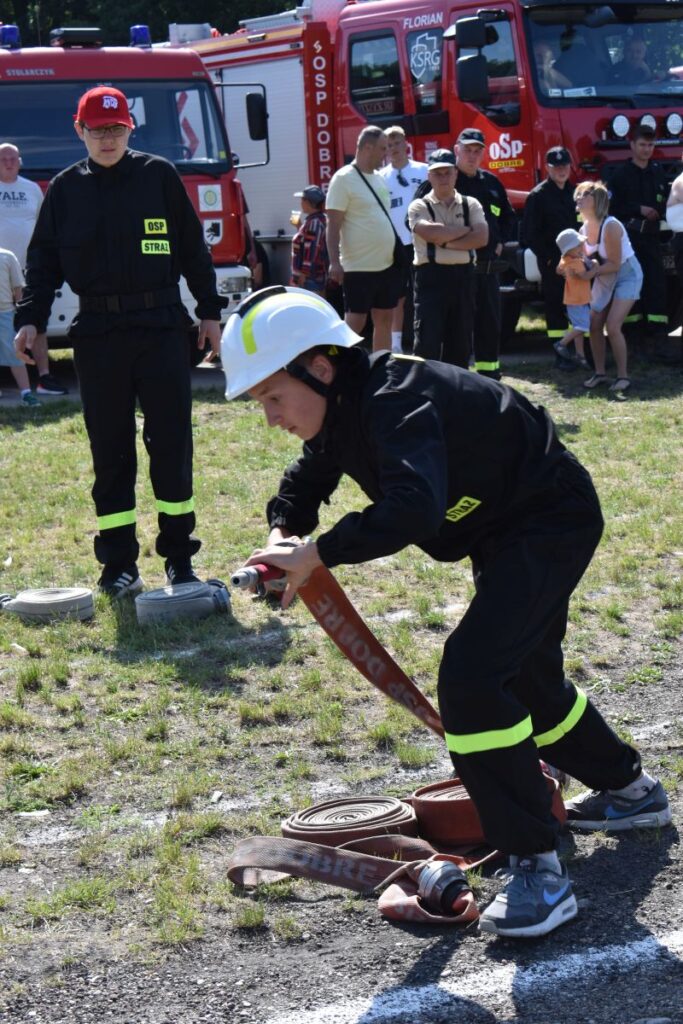 Na obrazku widocznym w odpowiednim urządzeniu obsługującym wąż strażacki podczas pokazu. W tle znajdują się inne strażacy oraz widzowie obok wozów strażackich.