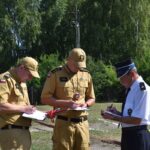 Na zewnątrz znajdują się trzy podstawowe elementy, które znajdują się w papierkowej roboty. Dwóch z nich ma na sobie beżowe mundury i czapki, podczas gdy trzeci ubrany jest w białą koszulę, czarne spodnie oraz czarną czapkę; w tle są drzewa i słoneczne niebo.