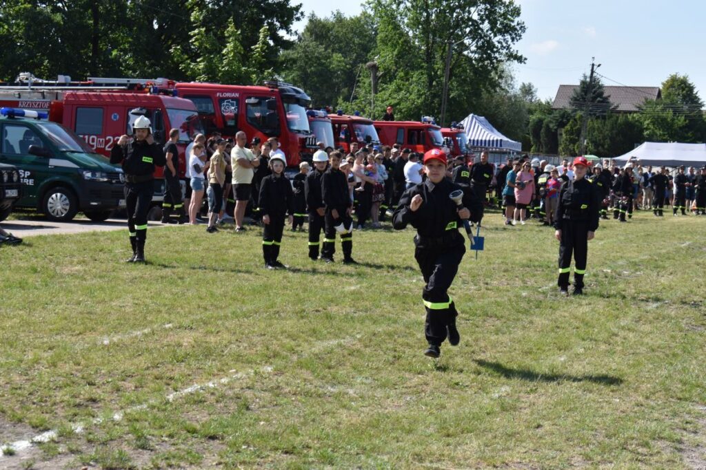 Na obrazku strażacy w mundurach uczestniczących w wydarzeniu plenerowym, na którym są symbole są wozy strażackie oraz zgromadzeni widzowie. Atmosfera specjalna, że jest pokazem lub ćwiczeniem jednostki pożarniczej.