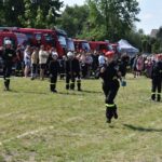 Na obrazku strażacy w mundurach uczestniczących w wydarzeniu plenerowym, na którym są symbole są wozy strażackie oraz zgromadzeni widzowie. Atmosfera specjalna, że jest pokazem lub ćwiczeniem jednostki pożarniczej.