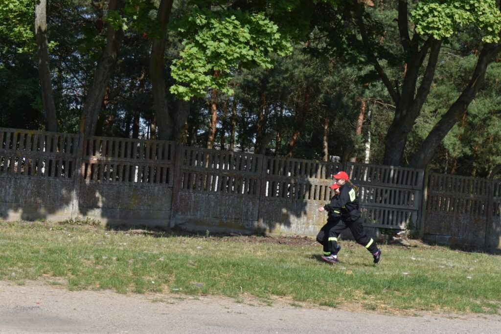 Na obrazku widoczna jest osoba ubrana w znaku strażackim i ograniczona czapka z daszkiem, która biegnie naprzód trawiastego terenu obok betonowego ogrodzenia. W tle znajduje się drzewo, które znajduje się w naturalnym środowisku.