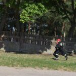 Na obrazku widoczna jest osoba ubrana w znaku strażackim i ograniczona czapka z daszkiem, która biegnie naprzód trawiastego terenu obok betonowego ogrodzenia. W tle znajduje się drzewo, które znajduje się w naturalnym środowisku.