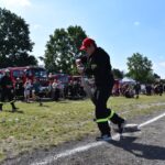 Na działaniu mundurze ciągnie wąż strażacki podczas zawodów, które odbywają się w słoneczny dzień. W tle widać, że istnieją grupy ludzkie oraz które są wozy strażackie.