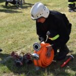 Na obrazku wodnym w ekwipunku kuca na trawie, zwijając pomarańczowy wąż obok kilku metalowych zaworów. W tle widoczni są ludzie i wózki dziecięce.