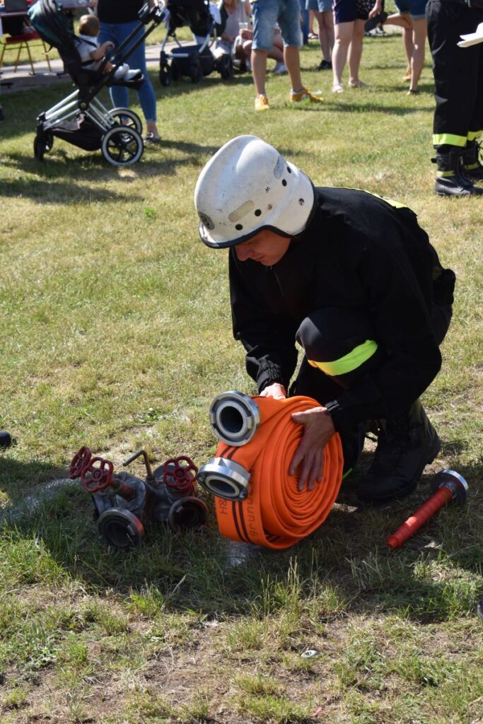 Na obrazku wodnym w ekwipunku kuca na trawie, zwijając pomarańczowy wąż obok kilku metalowych zaworów. W tle widoczni są ludzie i wózki dziecięce.