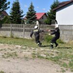 Na obrazku dwaj strażacy w mundurach biegnących obok ogrodzenia, z jednym szybko przychodzącym drugim. W tle widoczne są domy i drzewa.