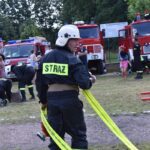 Na obrazku widocznym w ochronnym ubraniu z napisem „STRAŻ”, który trzyma żółty wąż pożarniczy. W tle znajdują się wozy strażackie oraz ludzie, którzy są częścią akcji.