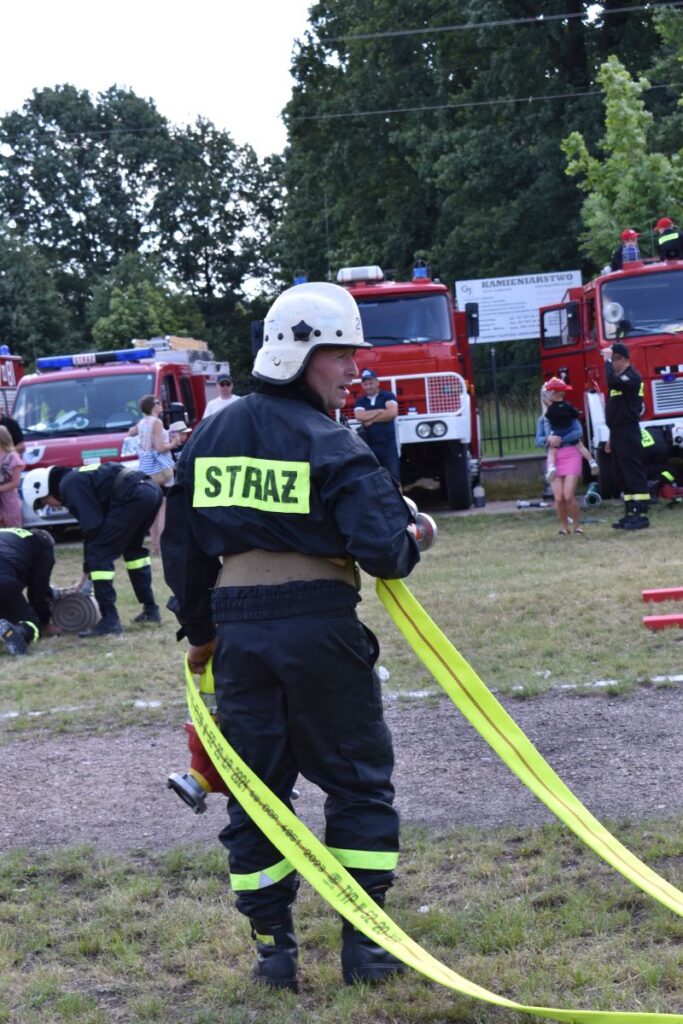 Na obrazku widocznym w ochronnym ubraniu z napisem „STRAŻ”, który trzyma żółty wąż pożarniczy. W tle znajdują się wozy strażackie oraz ludzie, którzy są częścią akcji.