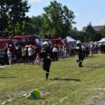 Na obrazku dwie osoby w ochronnym stroju biegnącym przez trawiastą polanę, a w tle widoczne są wozy strażackie oraz zgromadzeni obserwatorzy. Wydaje się, że scena stanowi akcję ratunkową lub działalność produkcyjną.