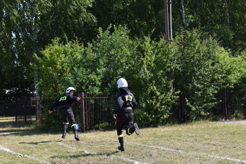 Na obrazku dwie osoby w czarnych kombinezonach ochronnych i białych kaskach z napisami „R2-D2” biegną po trawiastym polu w pobliżu bujnych drzew i ogrodzeń. Tło zielone stanowi krajobraz, wprowadzający spokojną, obniżoną okolicę.