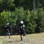 Na obrazku dwie osoby w czarnych kombinezonach ochronnych i białych kaskach z napisami „R2-D2” biegną po trawiastym polu w pobliżu bujnych drzew i ogrodzeń. Tło zielone stanowi krajobraz, wprowadzający spokojną, obniżoną okolicę.