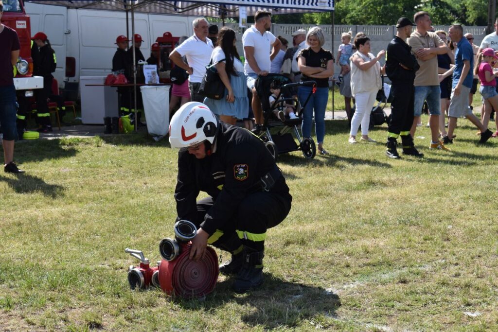 Strażak w mundurze reguluje wąż strażacki na trawienie podczas wydarzeń publicznych. W tle widoczni są ludzie, którzy obserwują i uczestniczą, a także namiot i sprzęt strażacki.