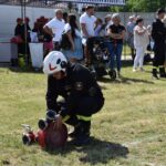 Strażak w mundurze reguluje wąż strażacki na trawienie podczas wydarzeń publicznych. W tle widoczni są ludzie, którzy obserwują i uczestniczą, a także namiot i sprzęt strażacki.