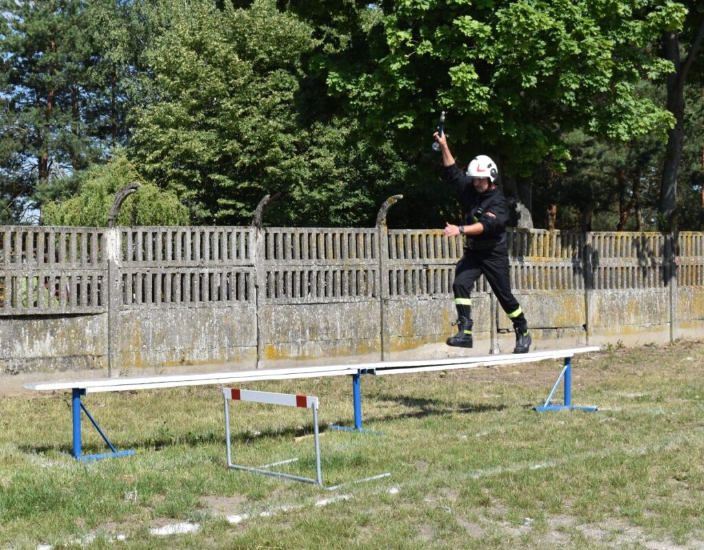 Na obrazie końcowym w mundurze balansuje na podniesionym belce podczas ćwiczeń szkoleniowych na świeżym powietrzu. W tle widać drzewo i ogrodzenie.