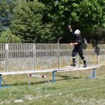 Na obrazie końcowym w mundurze balansuje na podniesionym belce podczas ćwiczeń szkoleniowych na świeżym powietrzu. W tle widać drzewo i ogrodzenie.