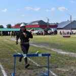 Na obrazku ochronnym w mundurze przeskakuje przez przeszkodę podczas zabiegu na trawiastym polu. W tle widać innych uczestników oraz osoby obserwujące, które są obserwowane.
