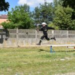 Strażak w podstawowym mundurze i hełmie jest uchwycony w powietrzu, przeskakując przez małą przeszkodę na trawiastej polanie. W tle widać betonowy płot oraz drzewo, powstające scenerie.