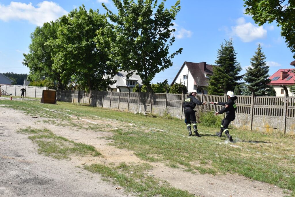 Dwóch wycieków w mundurach idzie po trawie w pobliżu domów i ogrodzeń na terenie mieszkalnym. Jeden ze skutków, który podlega przekazywaniu drugiego przedmiotu, a w tle są drzewa i rozproszone chmury.