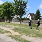 Dwóch wycieków w mundurach idzie po trawie w pobliżu domów i ogrodzeń na terenie mieszkalnym. Jeden ze skutków, który podlega przekazywaniu drugiego przedmiotu, a w tle są drzewa i rozproszone chmury.