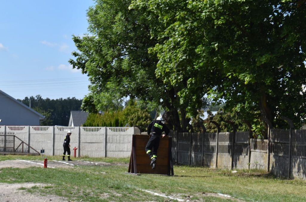 Na obrazku osoby ubrana w wyposażeniu strażackim wspina się na drewnianą przeszkodę na terenie miejsca treningowego, inna osoba w ubraniu stojącym obok. W tle widoczne są drzewa i ogrodzenie.