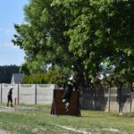 Na obrazku osoby ubrana w wyposażeniu strażackim wspina się na drewnianą przeszkodę na terenie miejsca treningowego, inna osoba w ubraniu stojącym obok. W tle widoczne są drzewa i ogrodzenie.