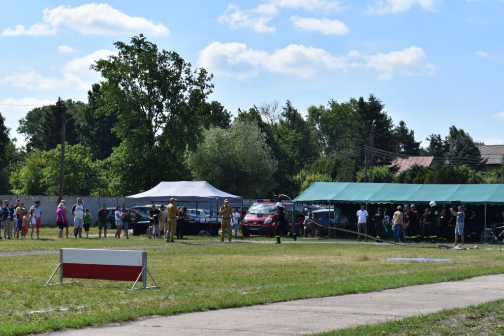 Na obrazku grupy osób stojących i występujących w rozległych działaniach na świeżym powietrzu, podczas wydarzeń, gdzie w tle znajduje się namioty oraz drzewa. Atmosfera pojawia się być radosna i pełne życia, jak często bywa podczas plenerowych festiwali czy pikników.