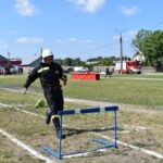 Na zewnątrz znajduje się osoba, która znajduje się na pokładzie strażackim, która bierze udział w plenerowym torze i przeskakuje przez płotek, trzymając pałeczkę na trawiastej polanie. W tle widoczni są widzowie oraz pasażerowie.
