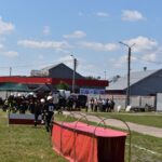 Na obrazku strażacy podzieleni na urządzenia na świeżym powietrzu, używając sprzętu i czerwonych tuneli na trawiastym polu. W tle widoczni są widzowie oraz inne osoby znajdujące się w pobliżu budynków.